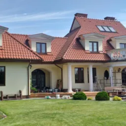 Elewacja domu z czerwonym dachem w trakcie malowania. Widoczne rusztowania i osoba pracująca na balkonie. Na trawniku narzędzia malarskie.