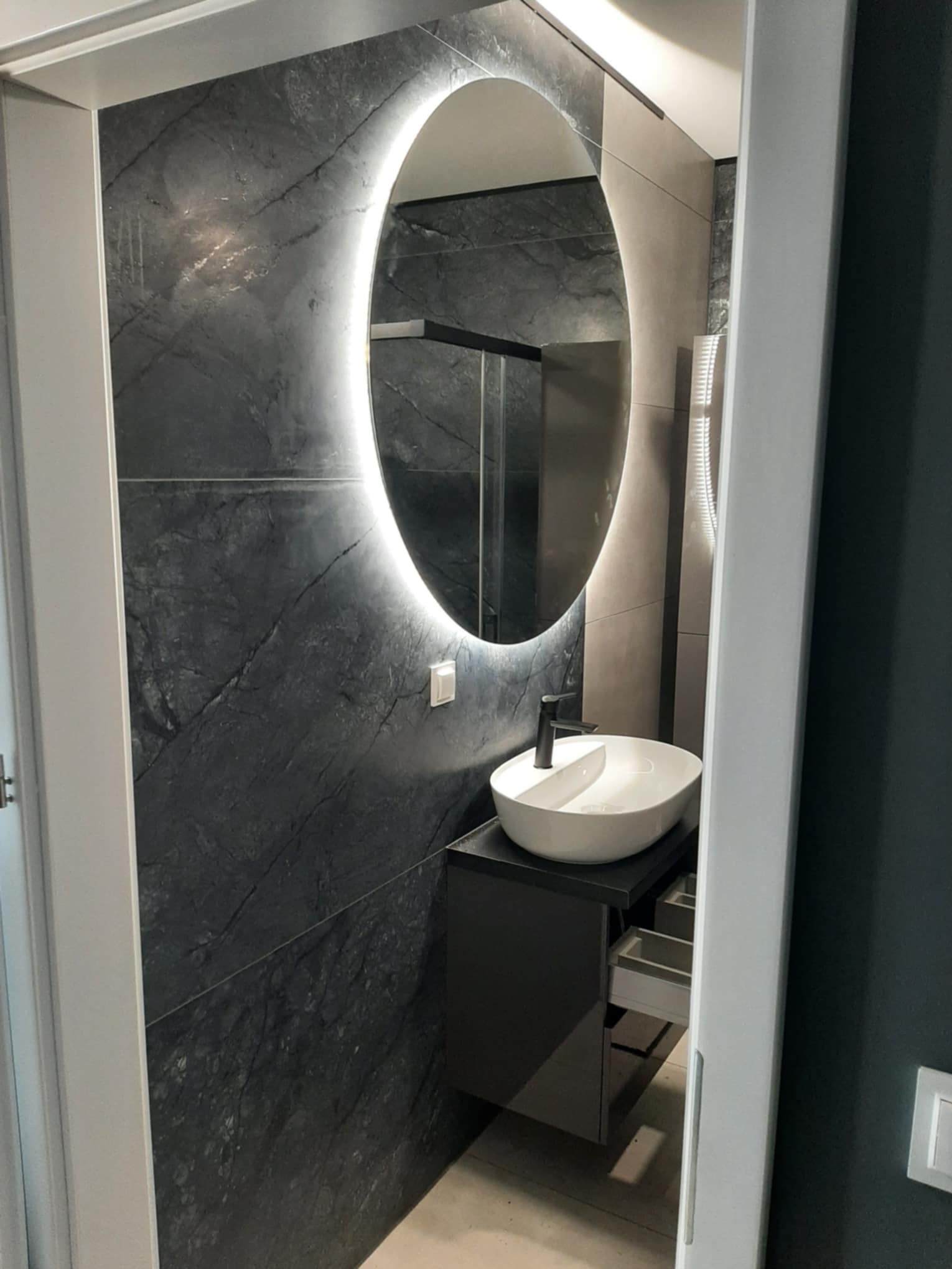 Nowoczesna łazienka z owalnym lustrem LED, umywalką nablatową i ścianą wyłożoną płytkami imitującymi ciemny marmur. Minimalistyczny design i eleganckie wykończenie.