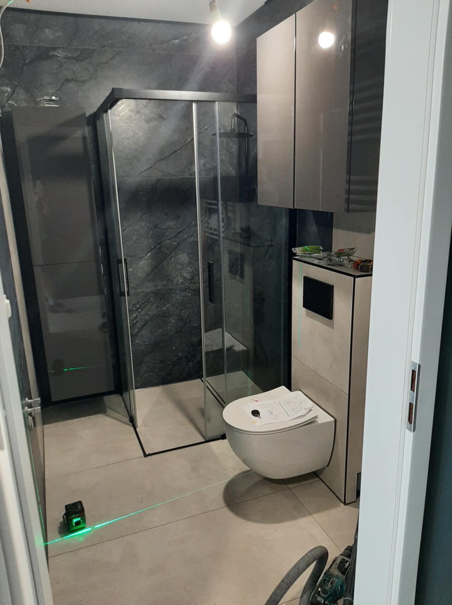 Nowoczesna łazienka z kabiną prysznicową, podwieszaną toaletą i szafkami w odcieniach szarości. Widoczny laser krzyżowy na podłodze.