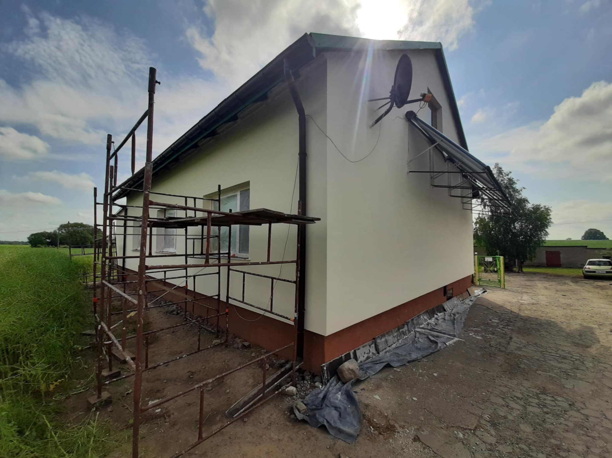 Dom po malowaniu elewacji, widoczne rusztowania i nowa, jasna kolorystyka ścian. Ciemna podmurówka i antena satelitarna na świeżo pomalowanej fasadzie.