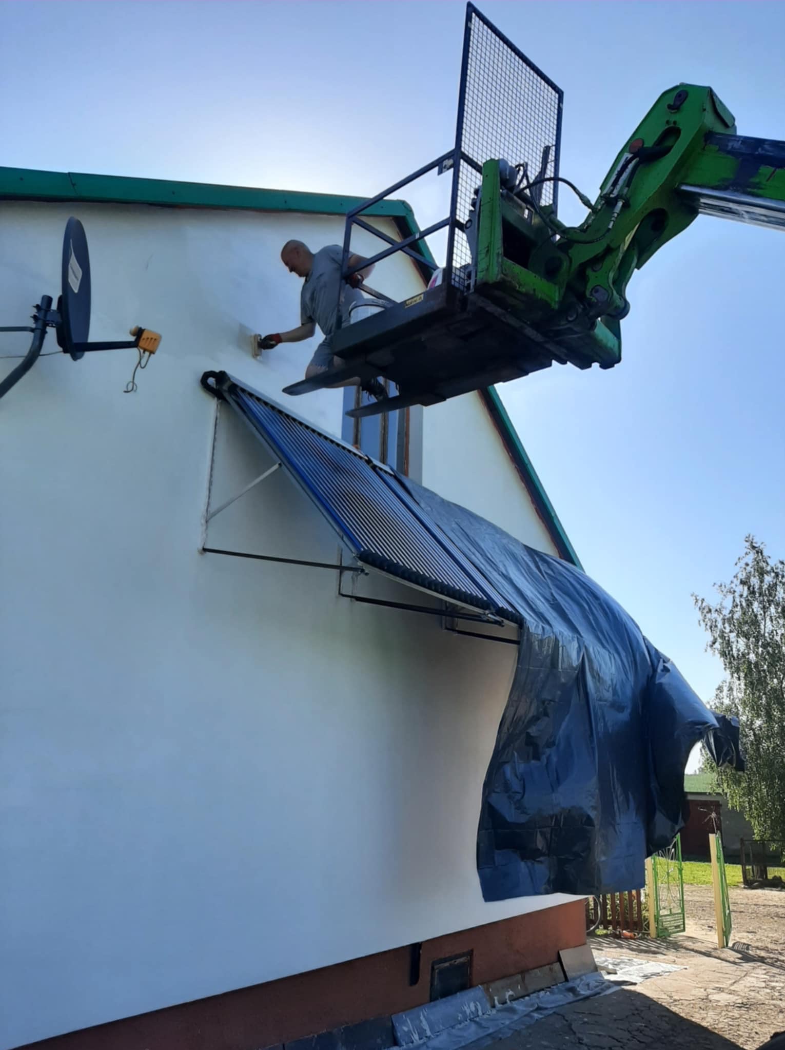 Pracownik na podnośniku maluje elewację domu. Widoczny fragment elewacji z osłoniętym kolektorem słonecznym. Słoneczny dzień.
