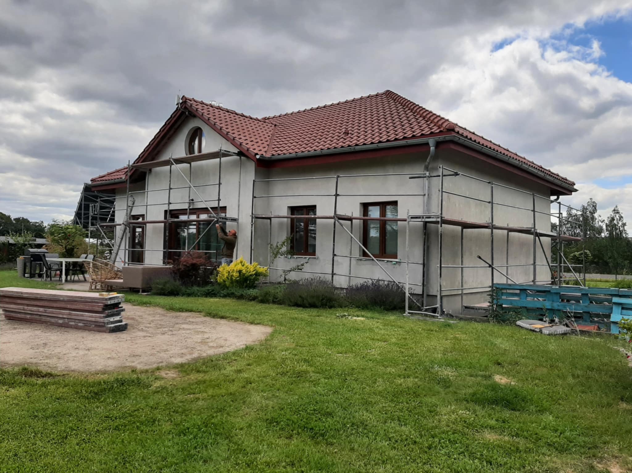 Dom jednorodzinny w trakcie prac elewacyjnych, otoczony rusztowaniem. Widoczne deski i narzędzia, sugerujące etap przygotowania do tynkowania. Pochmurne niebo w tle.