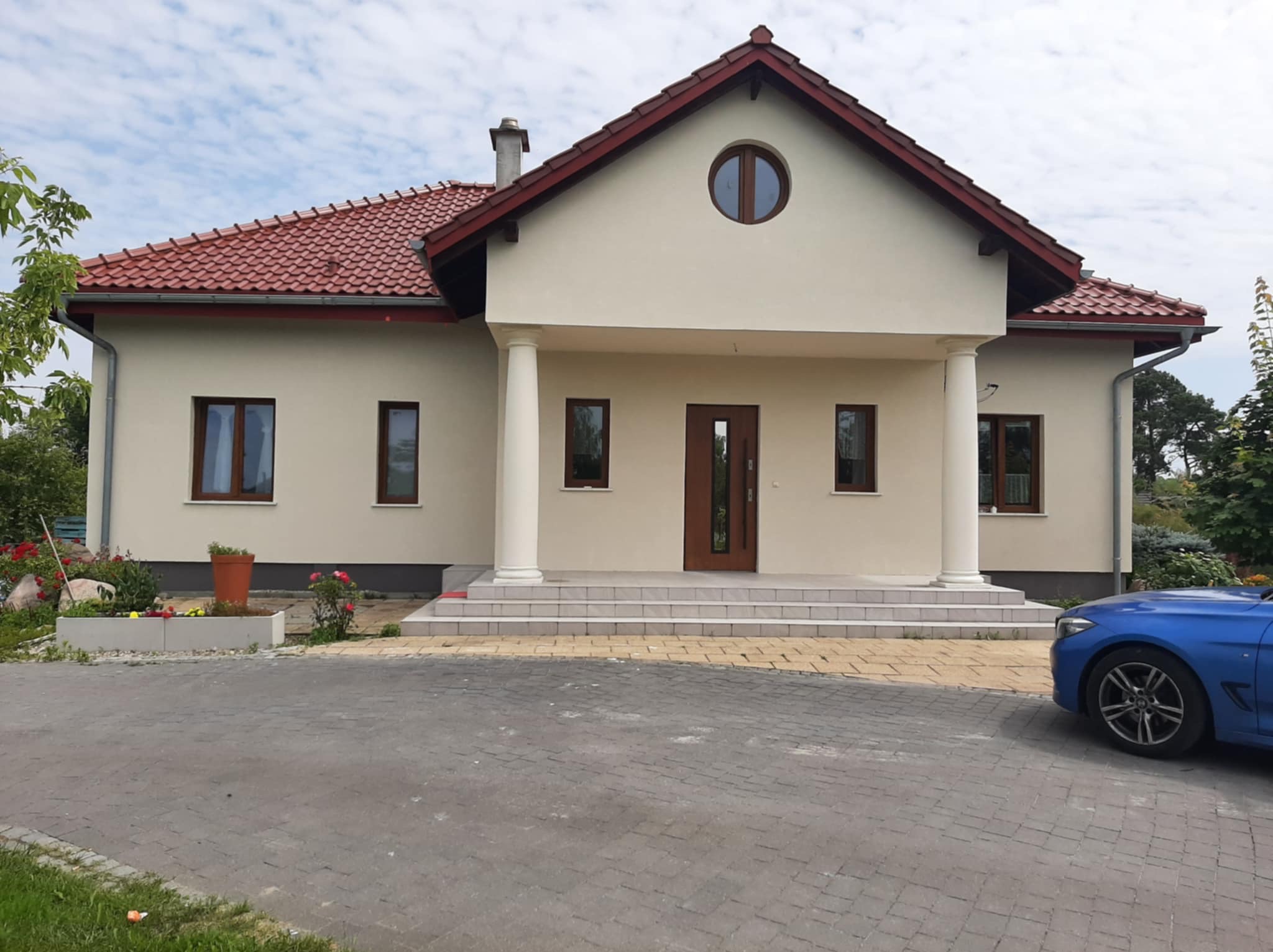 Jasny dom jednorodzinny z kremową elewacją, czerwoną dachówką i kolumnami wejściowymi, otoczony zielenią i brukowanym podjazdem. Widoczny fragment niebieskiego samochodu.