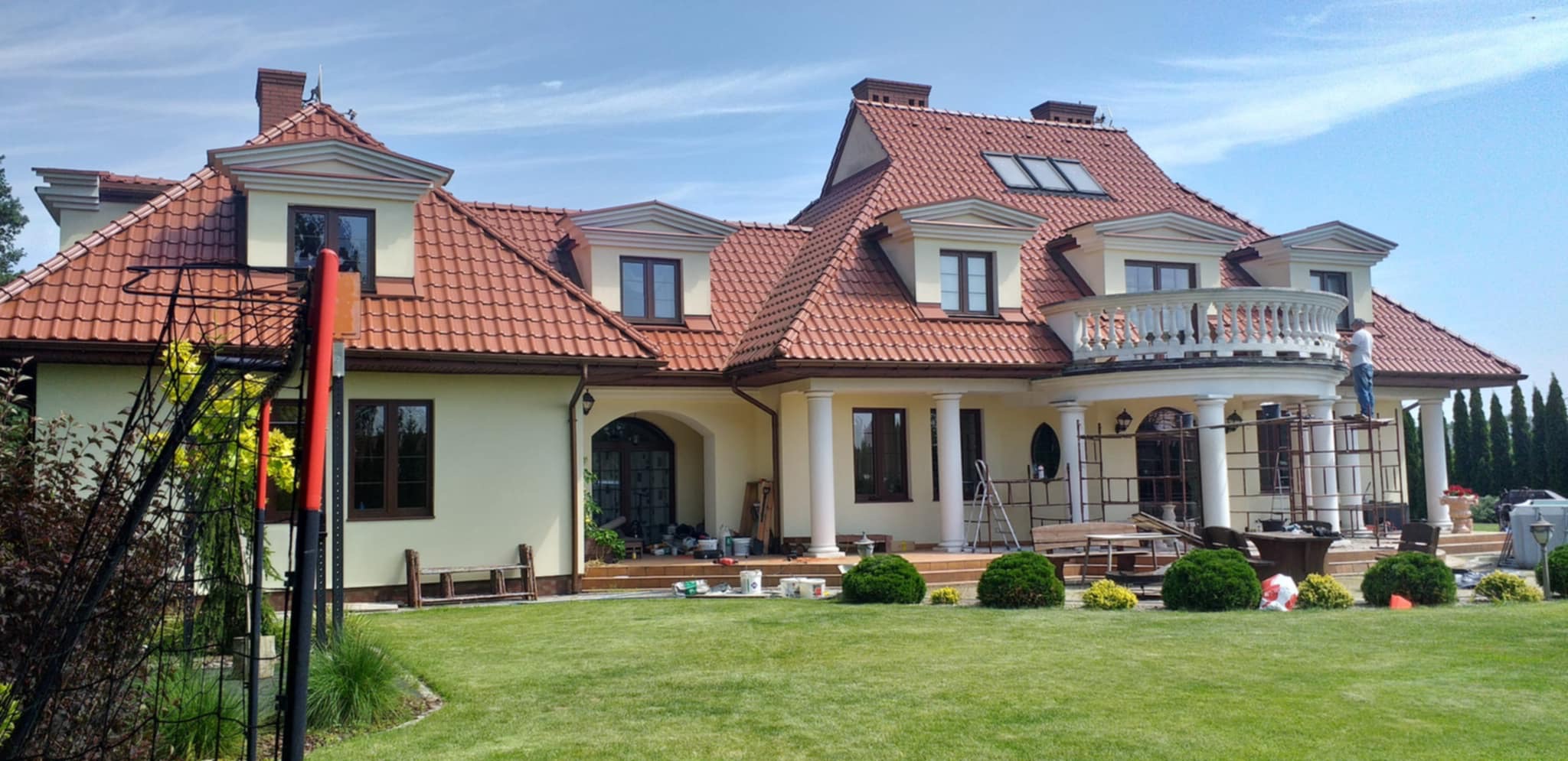Elewacja domu z czerwonym dachem w trakcie malowania. Widoczne rusztowania i osoba pracująca na balkonie. Na trawniku narzędzia malarskie.