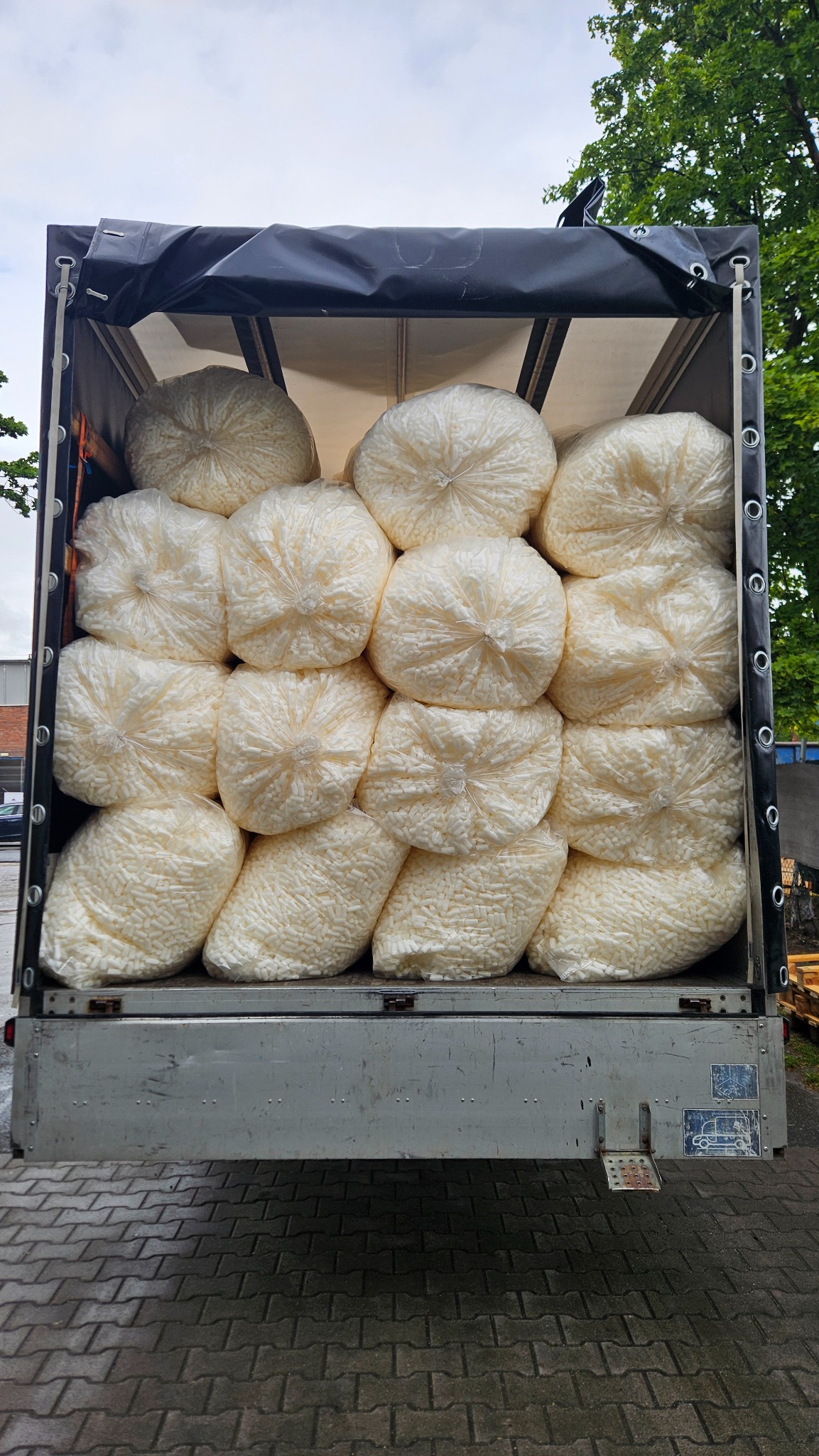 Przestrzeń ładunkowa busa wypełniona po brzegi białymi workami z wypełniaczem do paczek, widok z tyłu na otwartą pakę, kostka brukowa na dole.