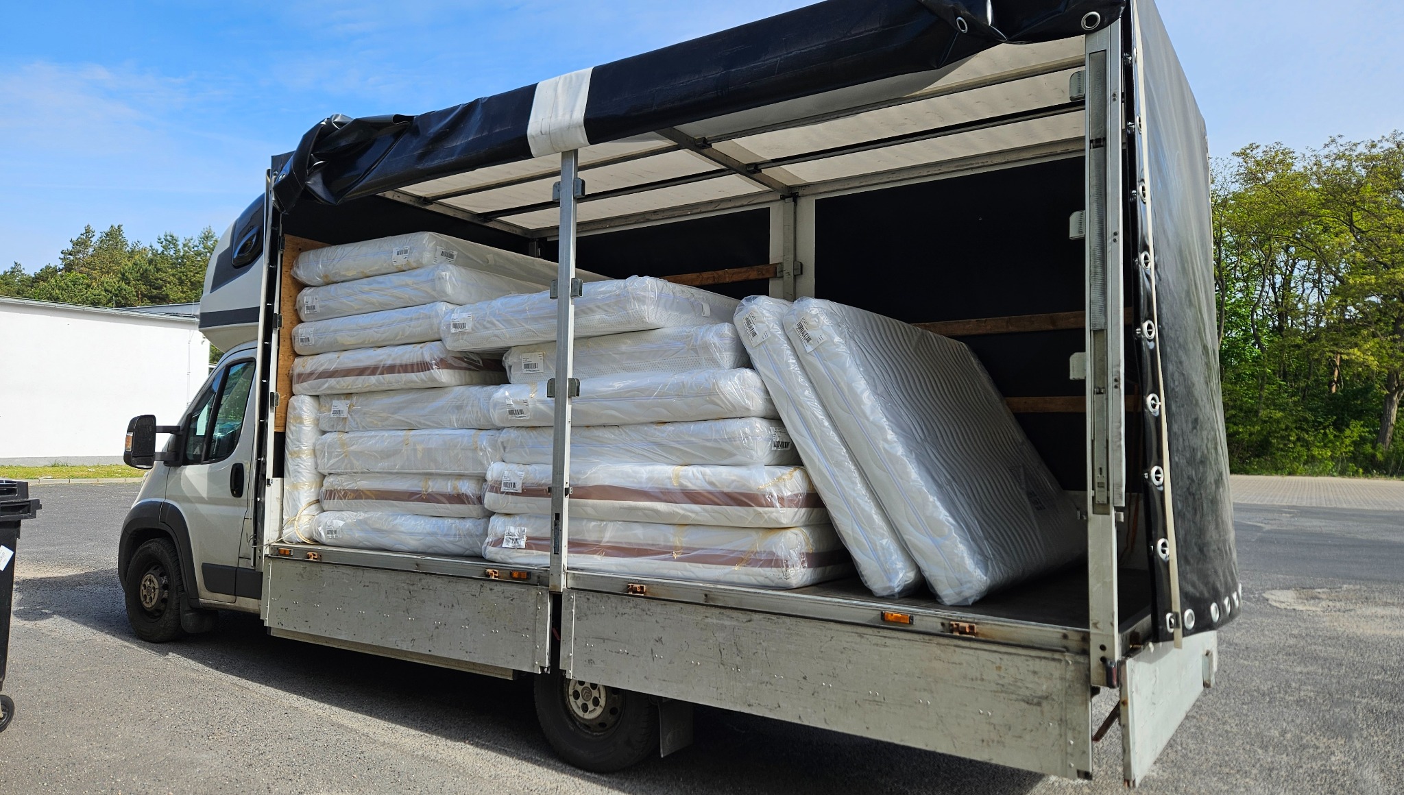 Biały bus z otwartą przestrzenią ładunkową, wypełnioną po brzegi zapakowanymi materacami. Częściowo czarna plandeka. Przewóz towarów.