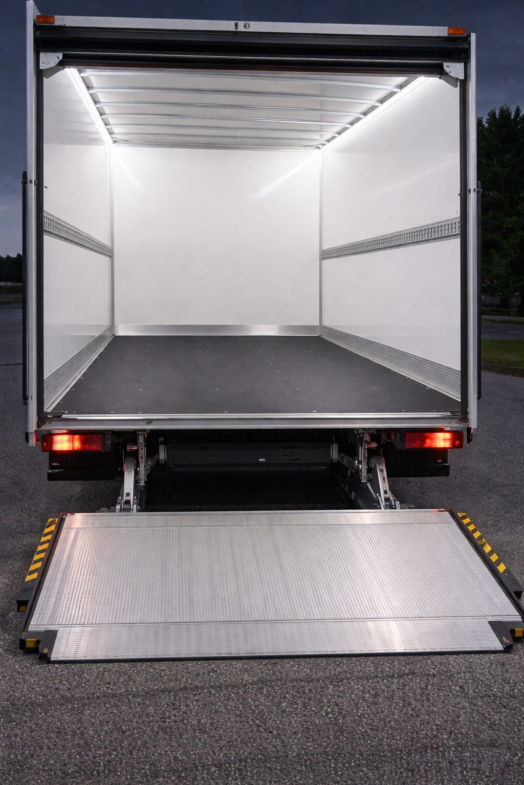 Wnętrze paki busa transportowego z otwartą, aluminiową rampą załadunkową. Widok z tyłu, oświetlenie LED wewnątrz. Pusta przestrzeń ładunkowa gotowa do transportu.