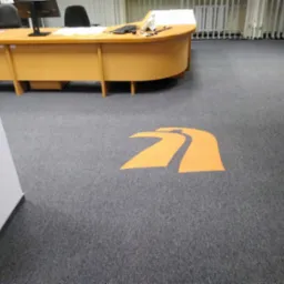 Szara wykładzina dywanowa w biurze z pomarańczowym logo firmy w kształcie strzałki, widok na biurko i grzejnik.