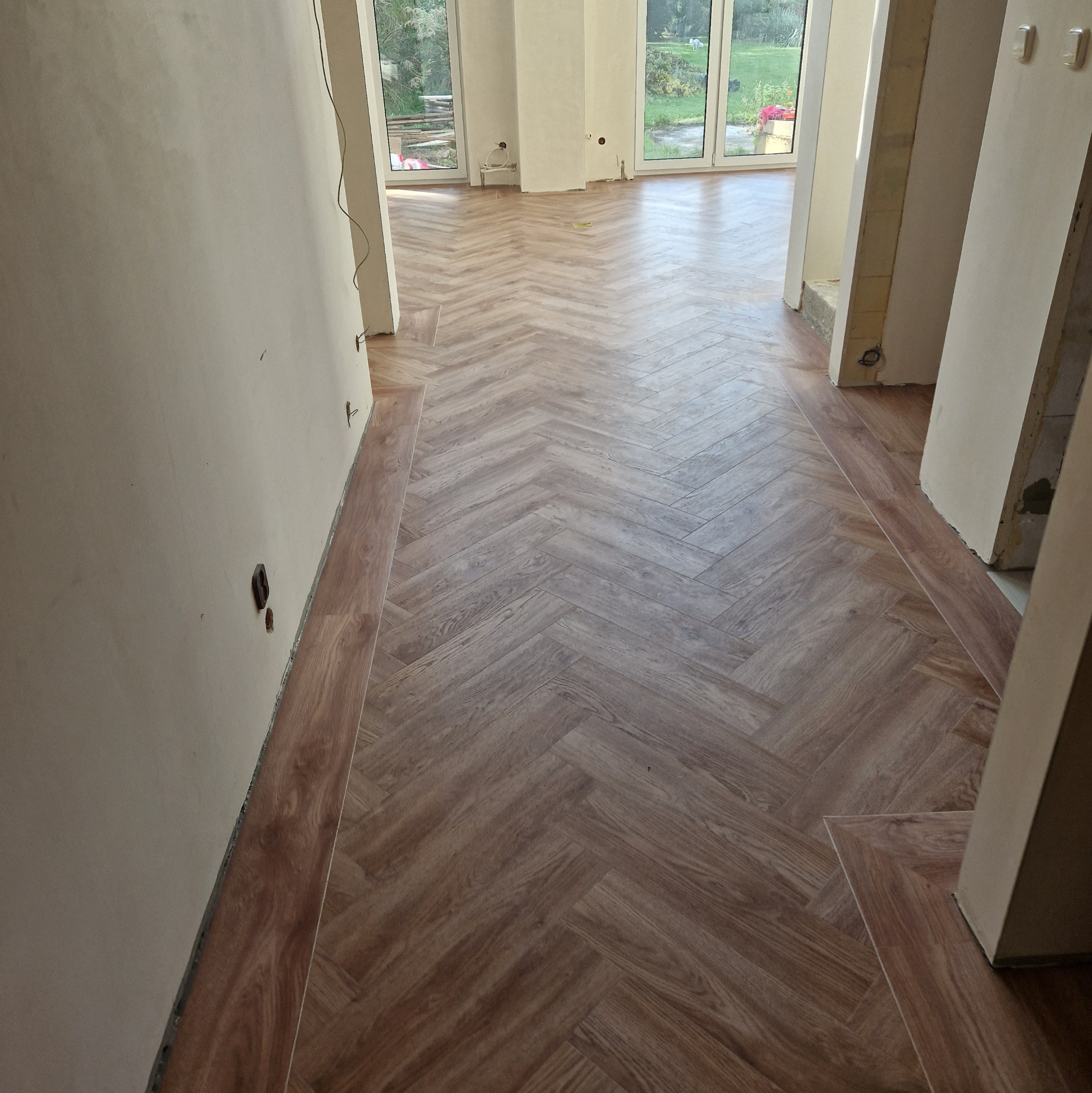 Układanie podłogi w jodełkę w jasnym pomieszczeniu z oknami, widoczne niedokończone ściany i listwy przypodłogowe.