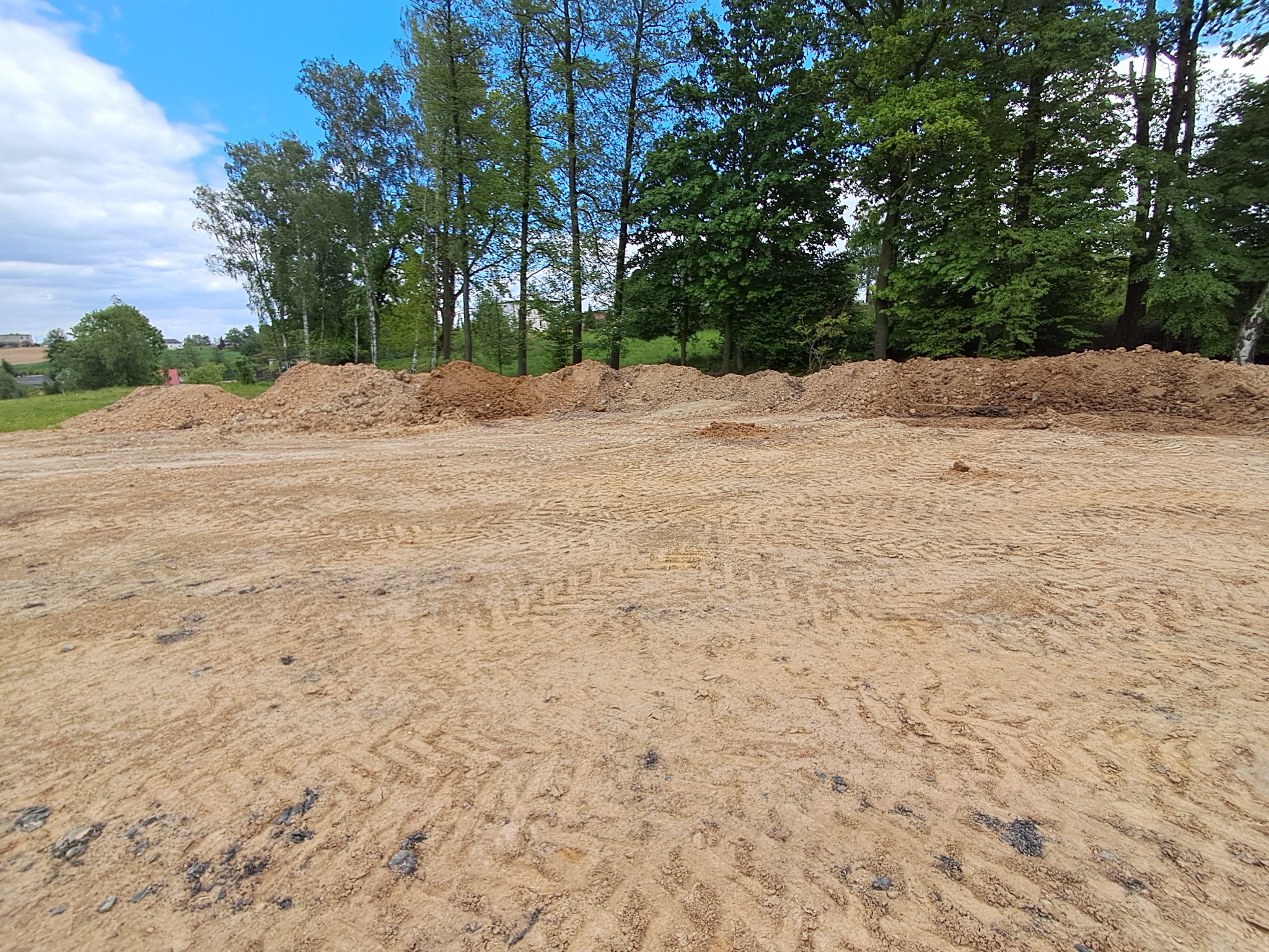 Wyrównany teren z widocznymi śladami opon, przygotowany pod dalsze prace budowlane. W tle drzewa i niebo. Niwelacja terenu w toku.