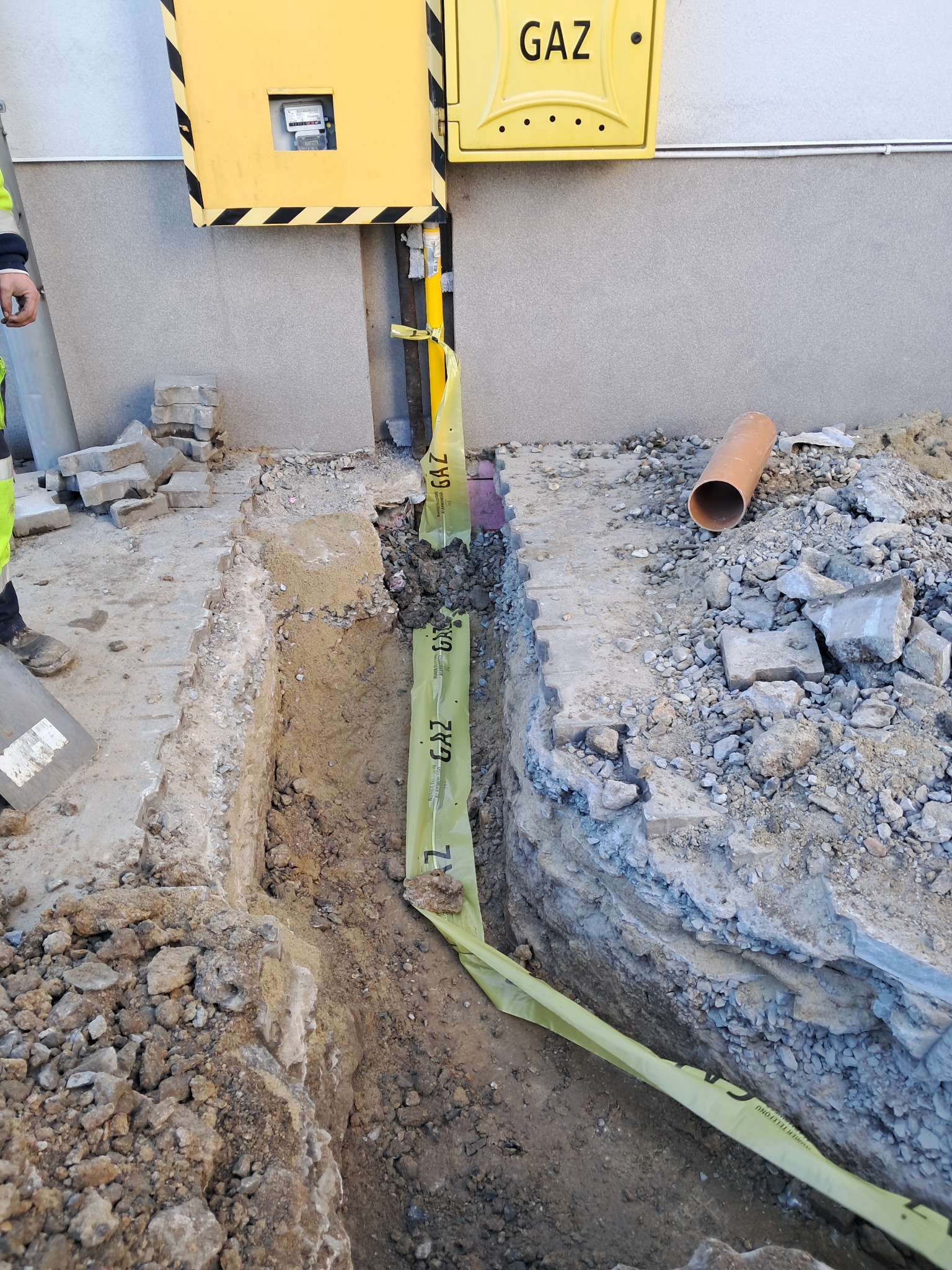 Wykopany wąski rów z żółtą taśmą ostrzegawczą GAZ, obok ściana budynku z żółtą skrzynką gazową. Widoczne gruz i rura. Prace ziemne przyłącza gazowego.