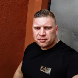 Portret mężczyzny w czarnej koszulce z logo EA7, z modną fryzurą, na tle pomarańczowej ściany i metalowej powierzchni. Wyraziste spojrzenie w obiektyw.