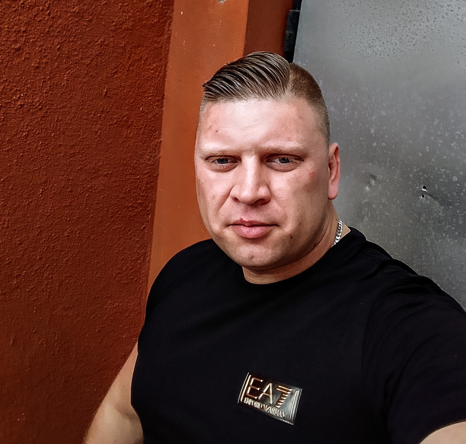 Portret mężczyzny w czarnej koszulce z logo EA7, z modną fryzurą, na tle pomarańczowej ściany i metalowej powierzchni. Wyraziste spojrzenie w obiektyw.