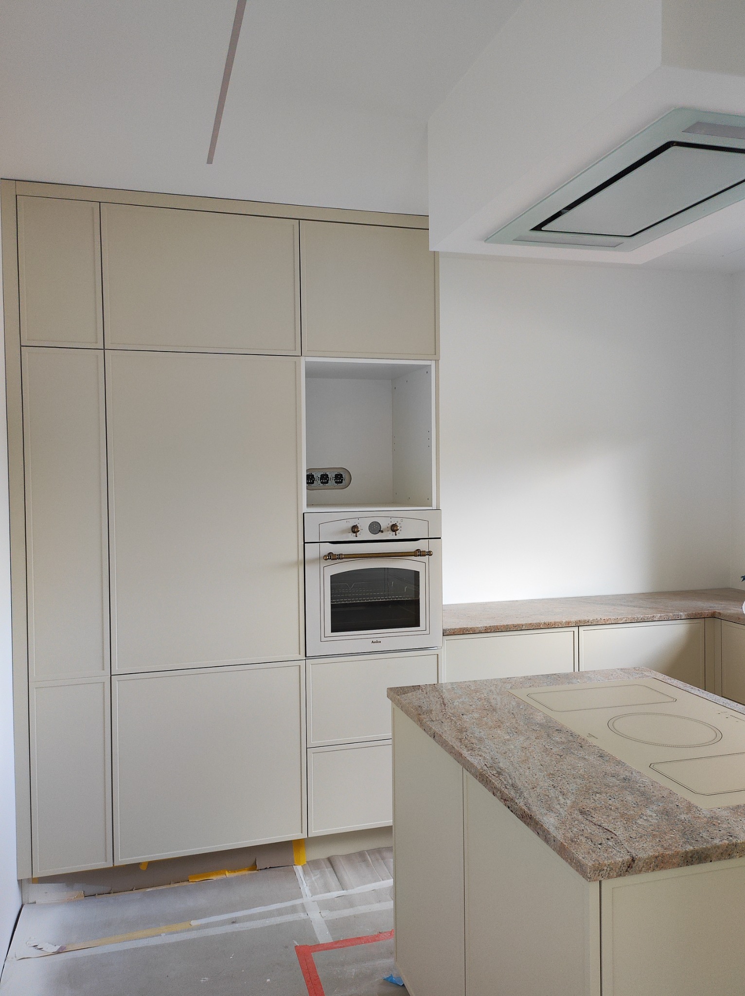 Jasna kuchnia w trakcie montażu: zabudowa meblowa z piekarnikiem Amica, wyspa z płytą grzewczą. Na podłodze widoczne ślady prac wykończeniowych.