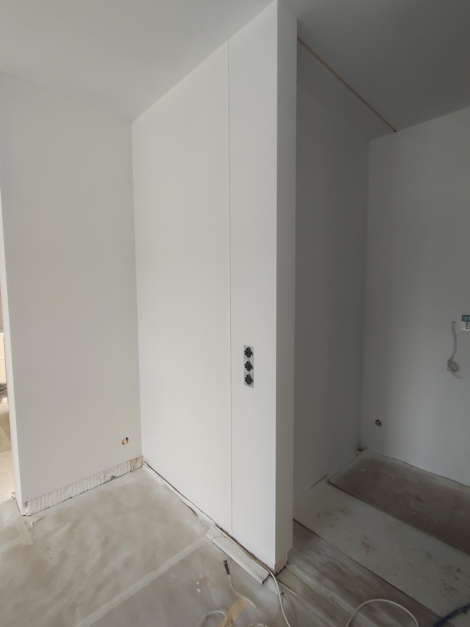 Białe, świeżo pomalowane wnętrze z widocznymi puszkami elektrycznymi i przygotowanym podłożem, sugerujące etap wykończeniowy. Minimalistyczny design.