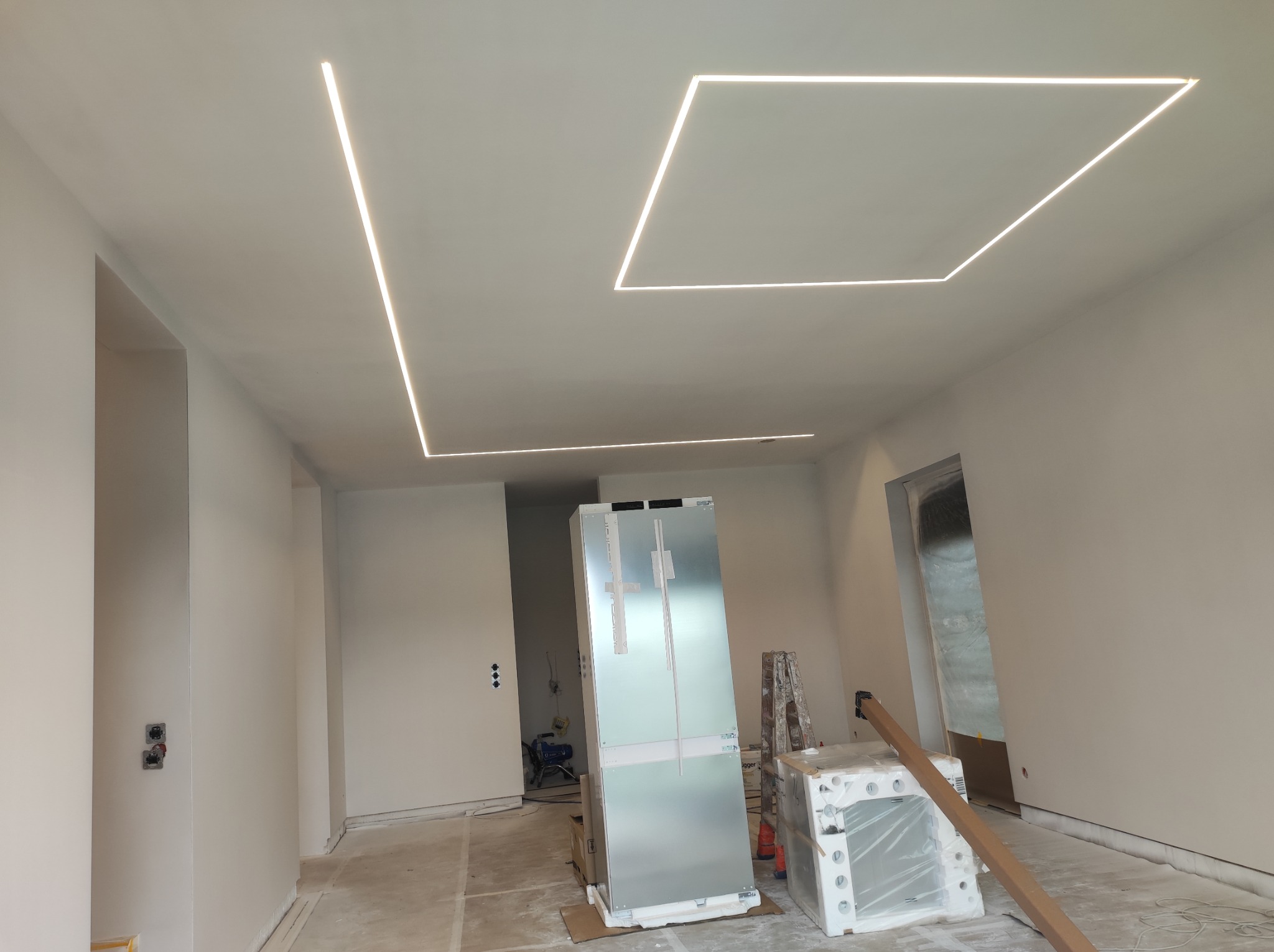 Białe wnętrze w trakcie wykończenia z geometrycznym oświetleniem LED na suficie. Na podłodze materiały budowlane i sprzęt. Widoczne ściany przygotowane do malowania.