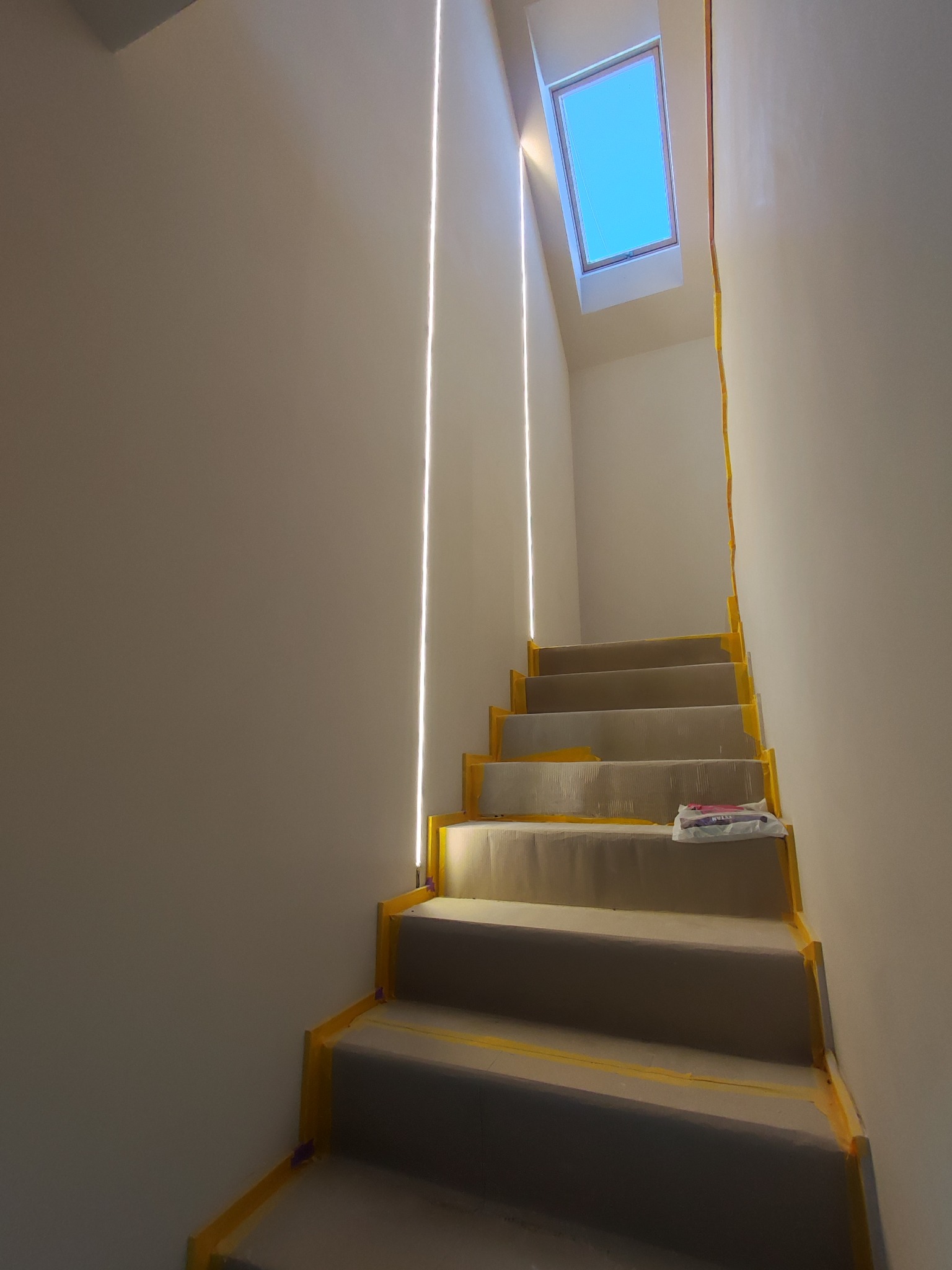 Schody po remoncie, zabezpieczone taśmą malarską, prowadzące na poddasze z oknem dachowym. Ściany pomalowane na jasny kolor, widoczne pionowe listwy oświetleniowe.