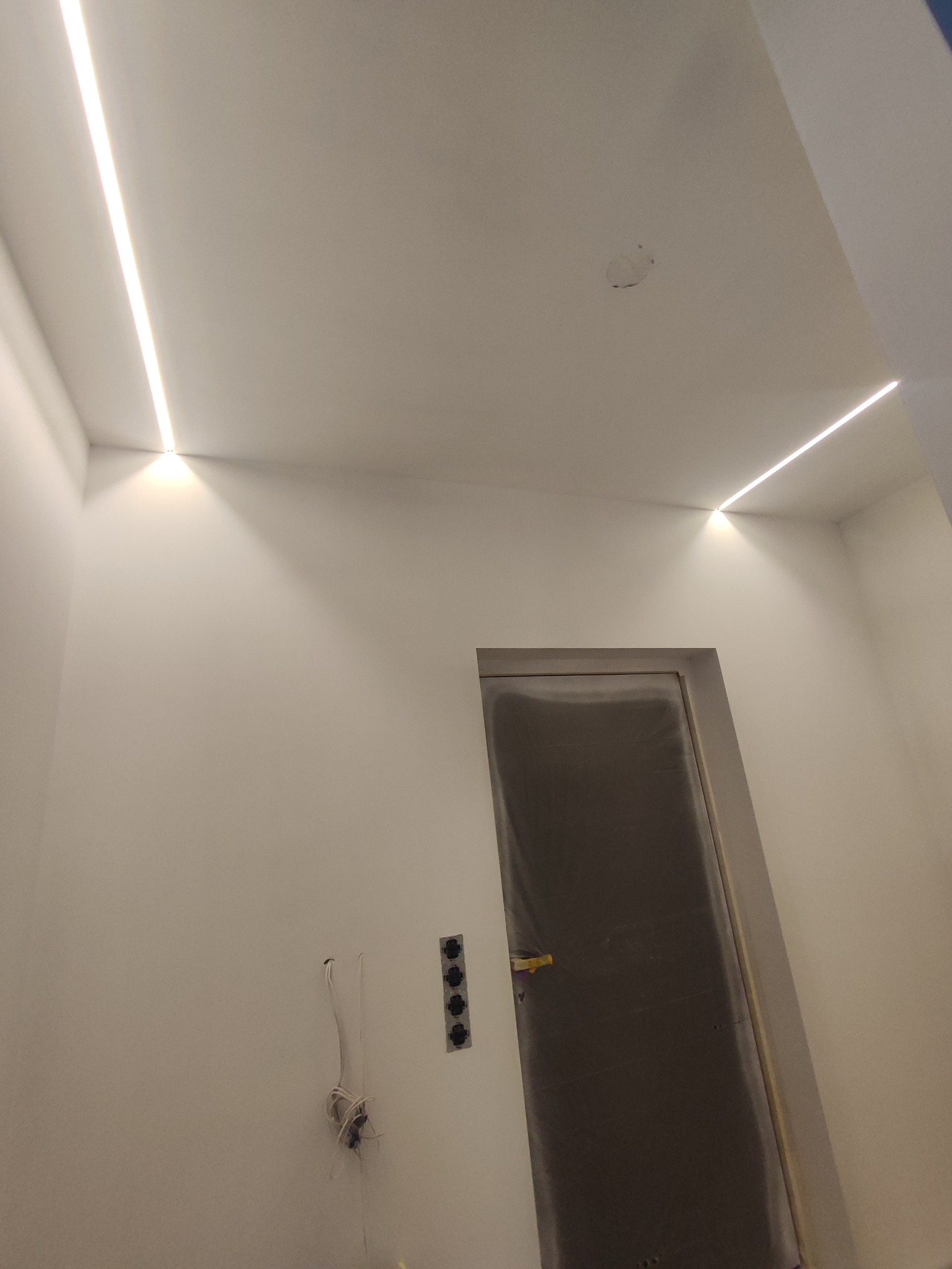 Minimalistyczne wnętrze w trakcie wykończenia: białe ściany, ukryte oświetlenie LED w suficie, zabezpieczone drzwi i przygotowane gniazdka elektryczne.