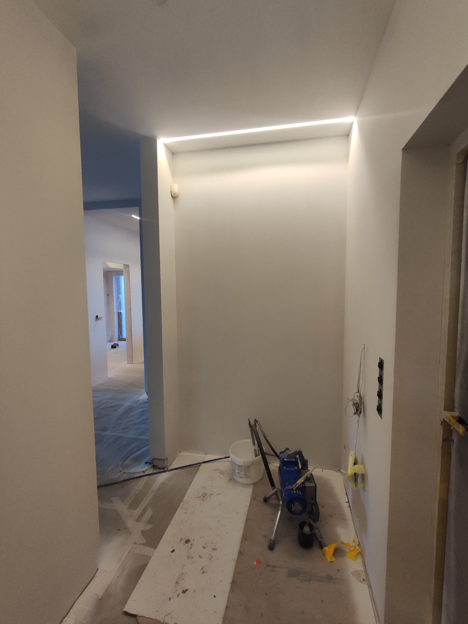 Świeżo pomalowane wnętrze z oświetleniem LED w suficie i sprzętem malarskim, gotowe do dalszych prac wykończeniowych. Widoczne gniazdka elektryczne.