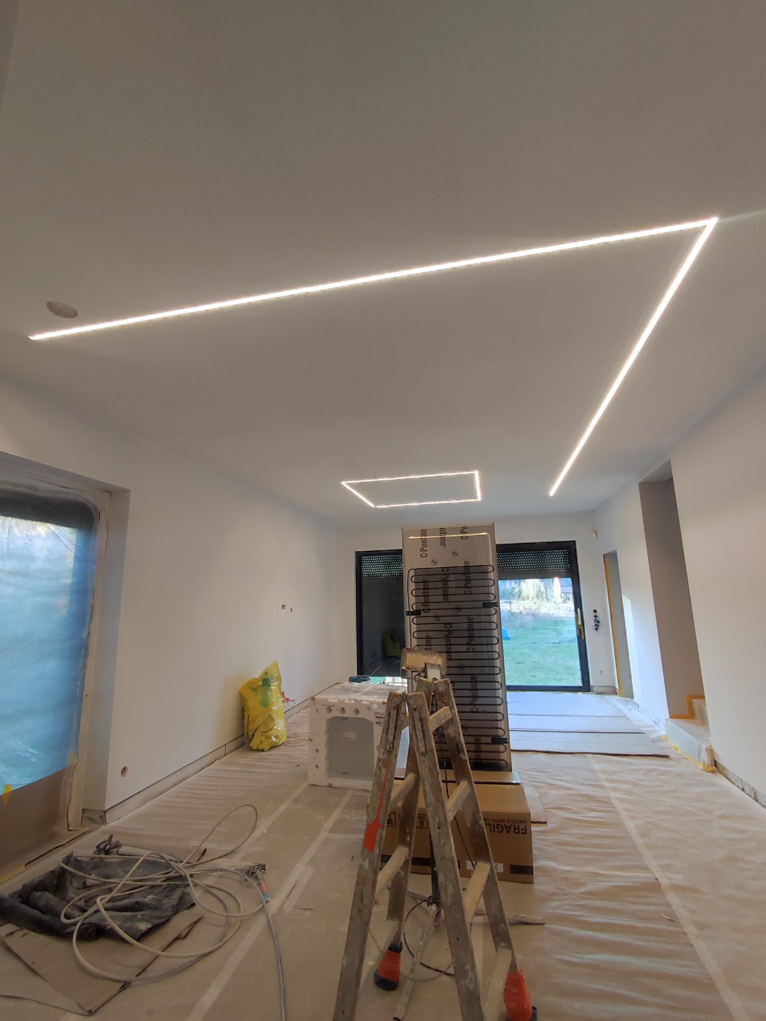 Wnętrze w trakcie wykończenia z zamontowanym oświetleniem LED w suficie. Widoczne narzędzia i materiały budowlane, zabezpieczone folią okna i podłoga.