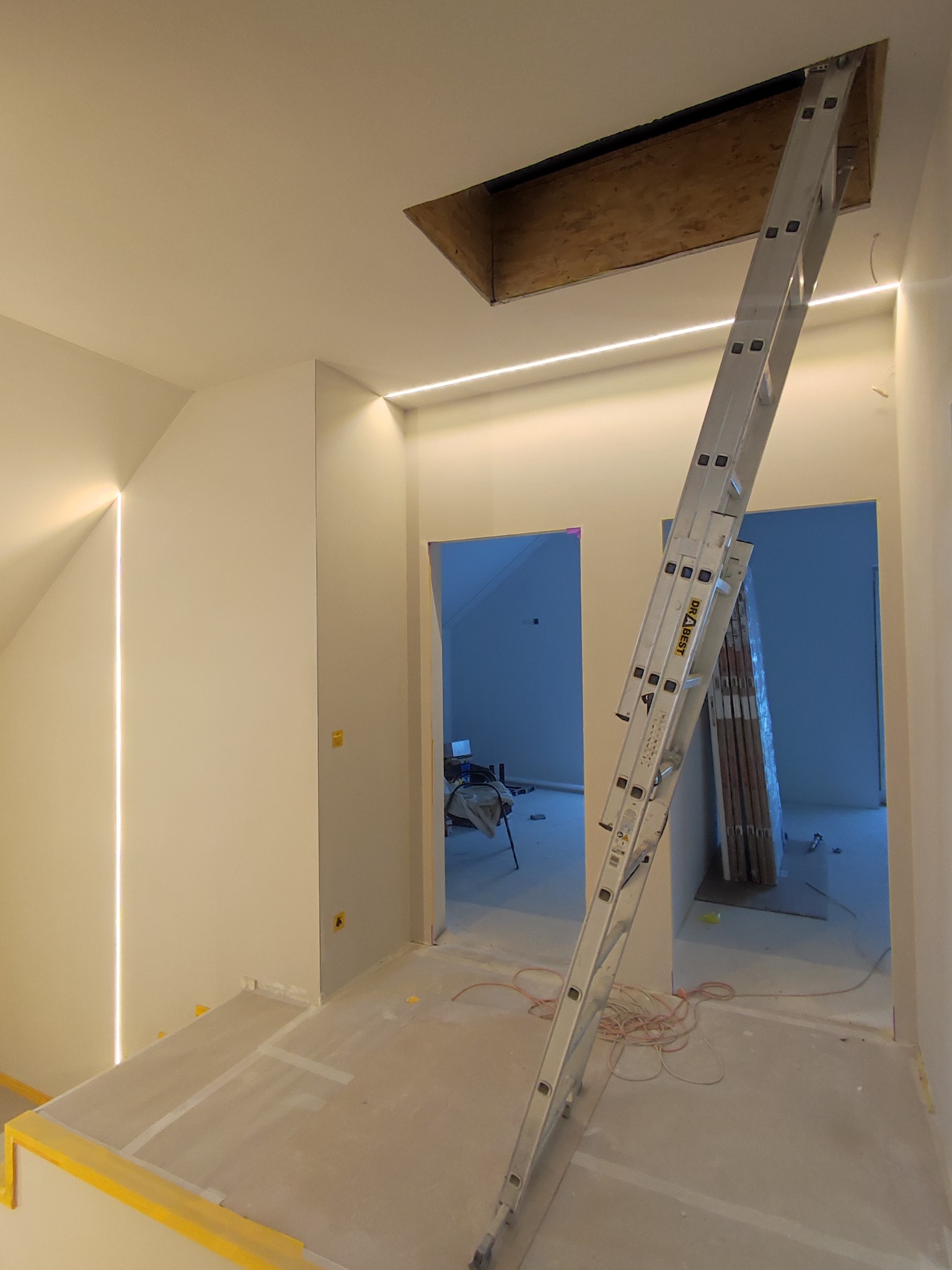 Wnętrze w Luboniu w trakcie prac wykończeniowych: widoczna drabina oparta o otwór w suficie, ściany przygotowane do malowania, oświetlenie ledowe wzdłuż ścian.