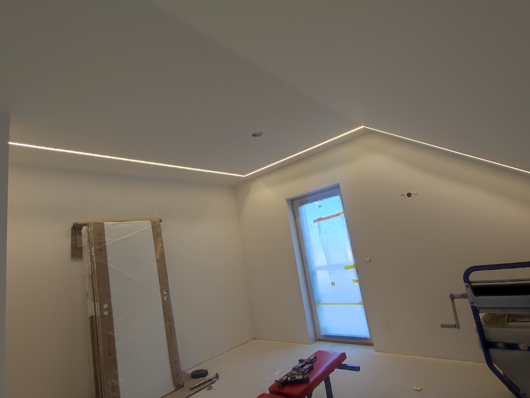 Pokój na poddaszu po remoncie: białe ściany, sufit z oświetleniem LED, drzwi w folii ochronnej i narzędzia. Etap wykończeniowy, widok z dołu.