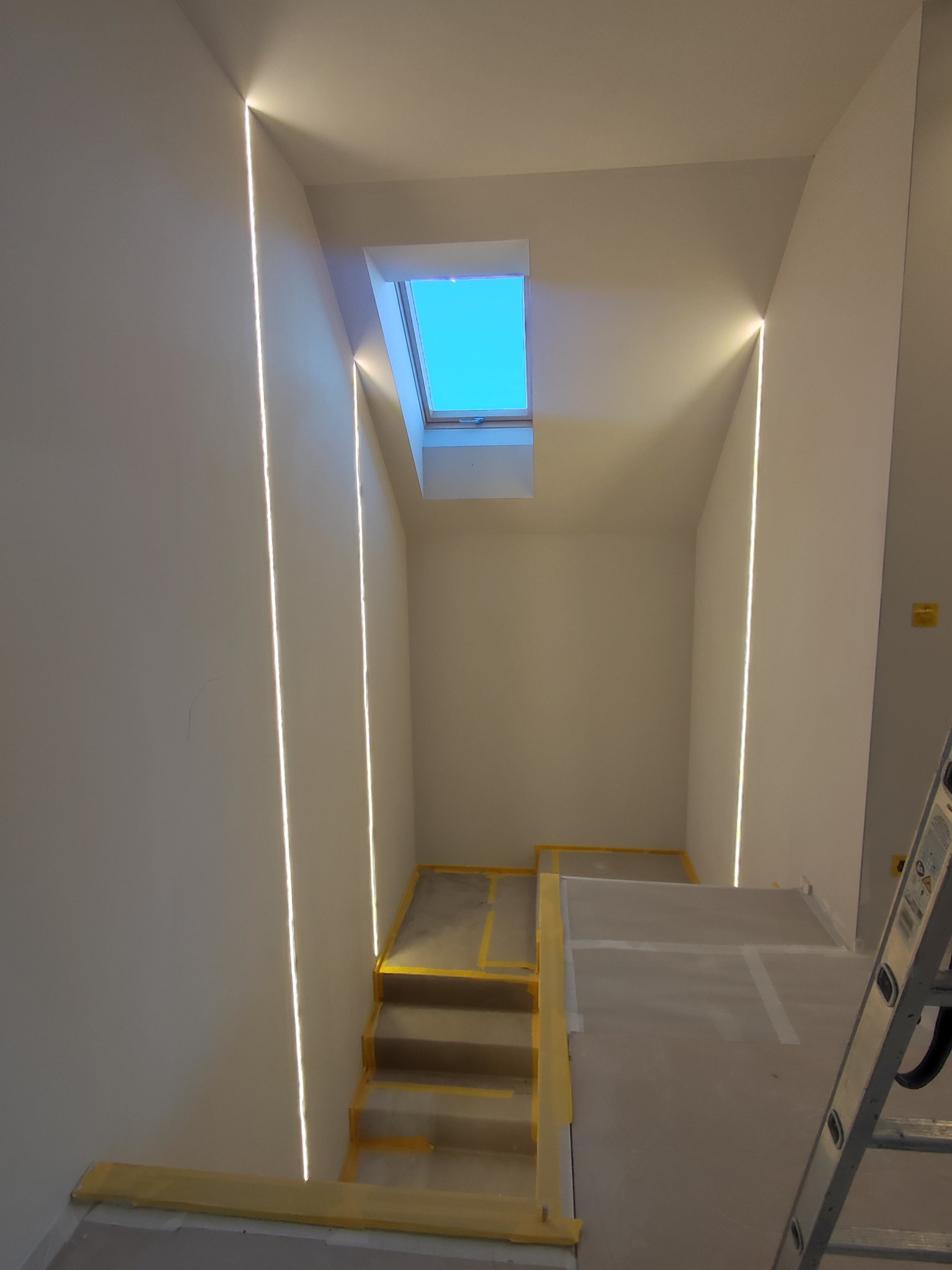 Schody w trakcie prac wykończeniowych, zabezpieczone taśmą malarską, z oknem dachowym i liniowym oświetleniem LED wzdłuż ścian.