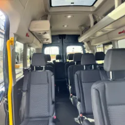 Wnętrze przestronnego busa pasażerskiego z rzędami czarnych foteli, ż&oacute;łtą poręczą przy wejściu i oknem dachowym. Widoczne detale jak młotek bezpieczeństwa i oświetlenie.