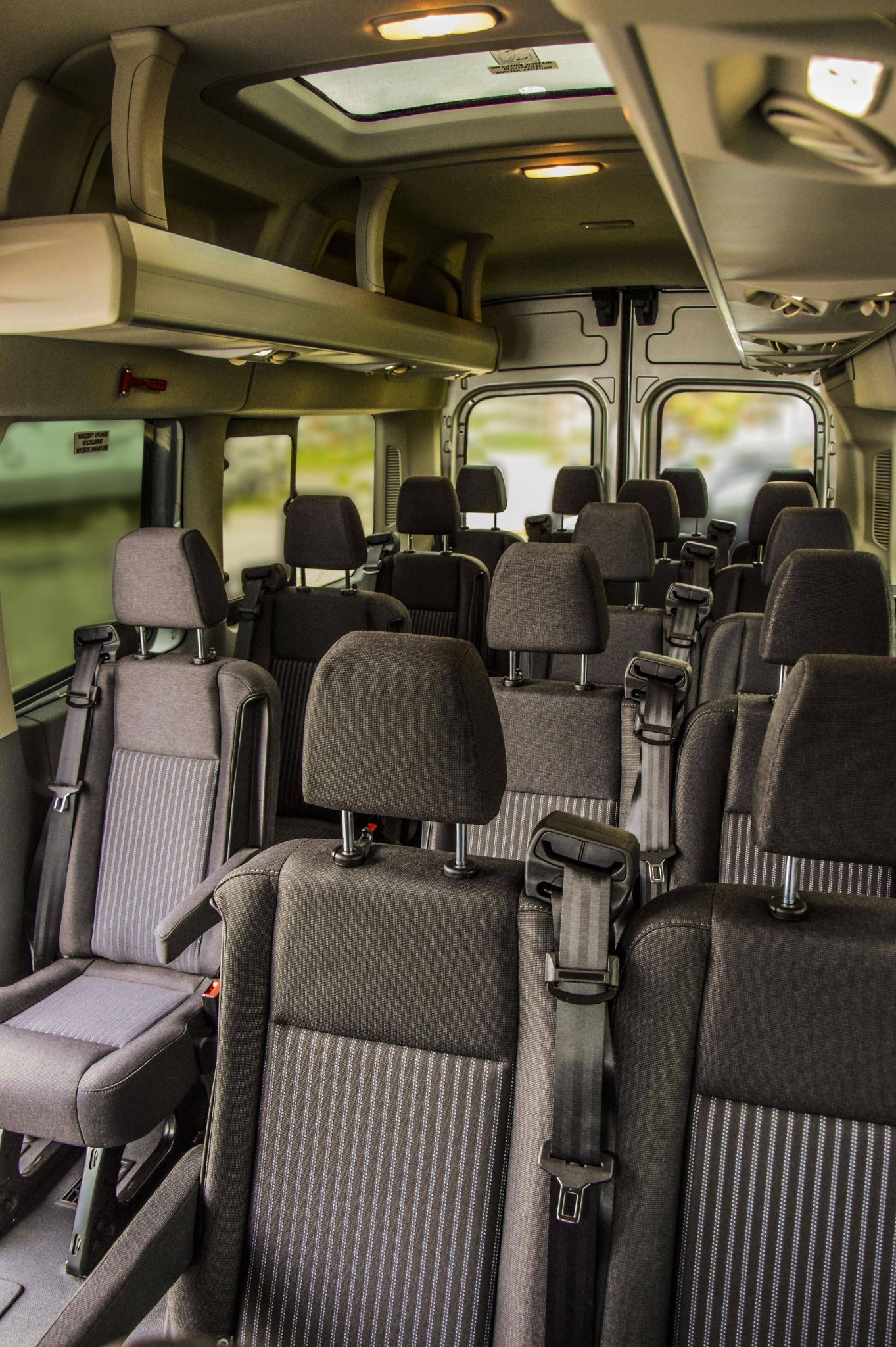 Wnętrze busa pasażerskiego z rzędami szarych foteli z pasami bezpieczeństwa, widok z tyłu kabiny, oświetlenie sufitowe i schowki nad głowami.