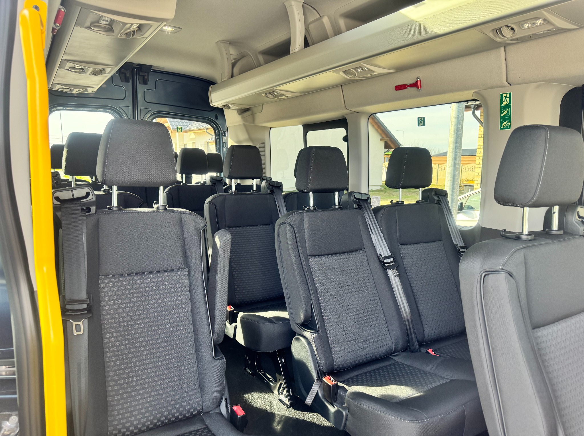 Wnętrze busa pasażerskiego z czarnymi fotelami i żółtą poręczą. Widoczne zagłówki, pasy bezpieczeństwa oraz okna z widokiem na budynki.