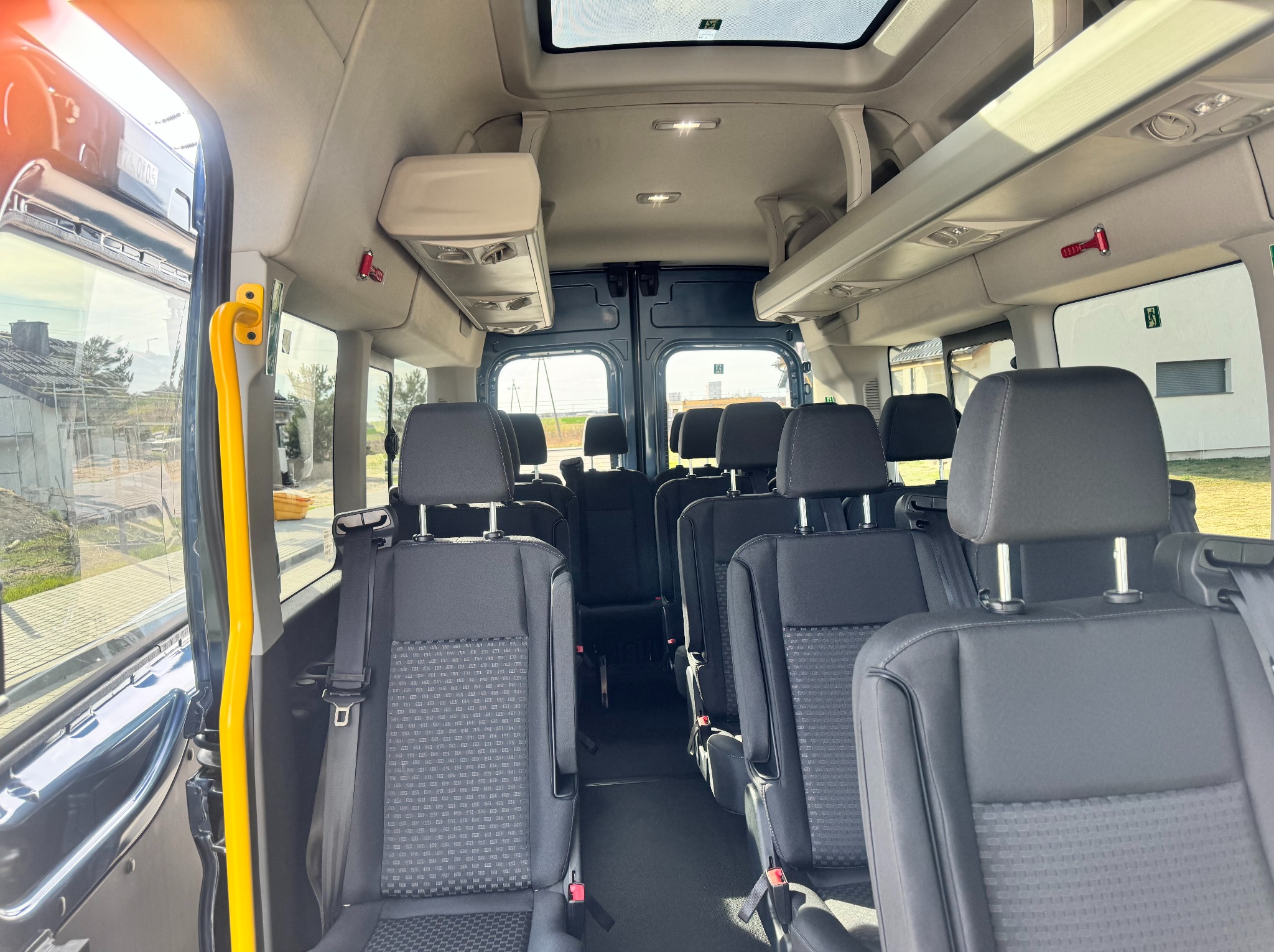 Wnętrze przestronnego busa pasażerskiego z rzędami czarnych foteli, żółtą poręczą przy wejściu i oknem dachowym. Widoczne detale jak młotek bezpieczeństwa i oświetlenie.