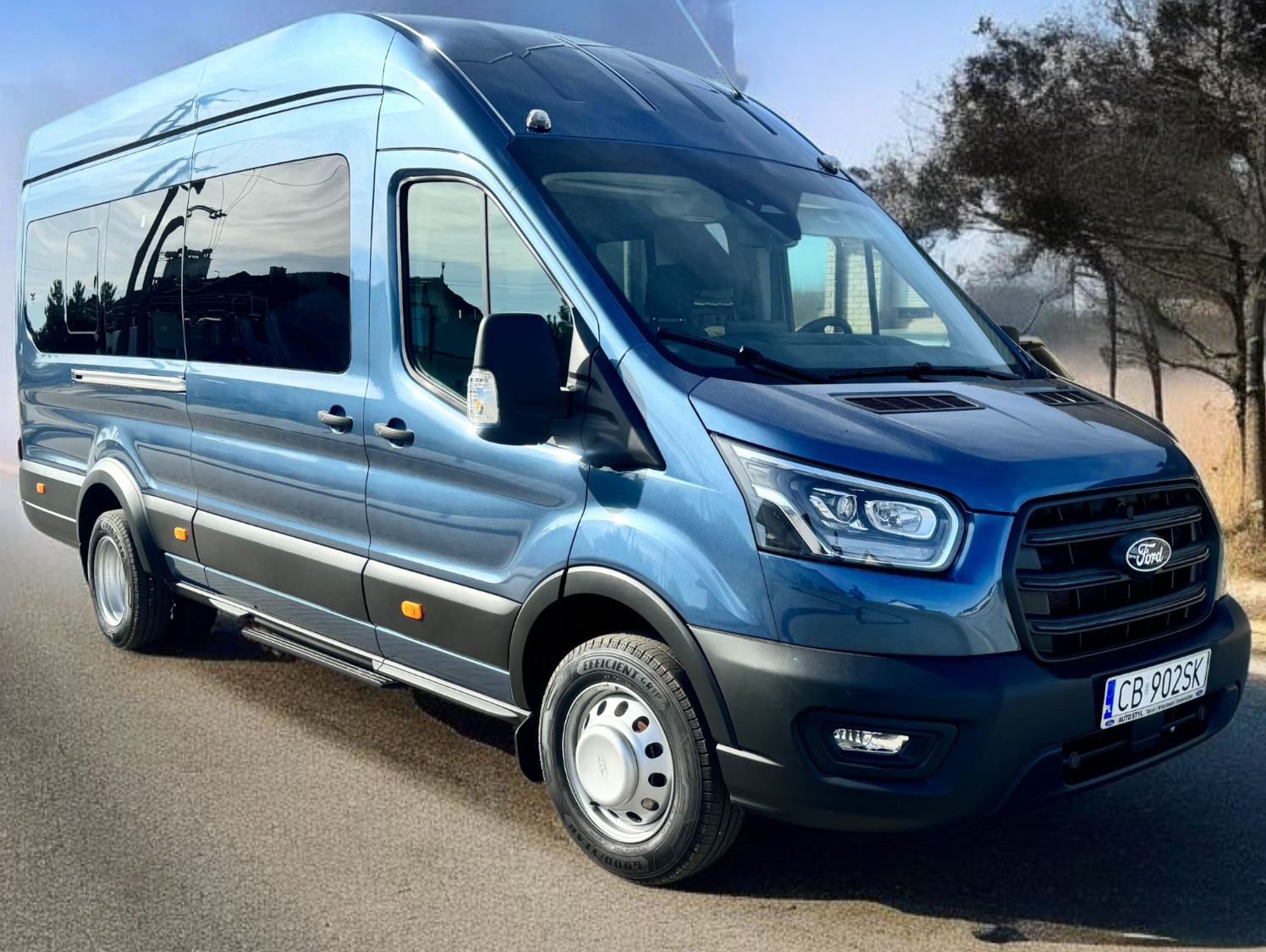 Błękitny, lśniący Ford Transit na asfalcie, z widocznym fragmentem tablicy rejestracyjnej i odblaskami w oknach, na tle drzew bez liści.