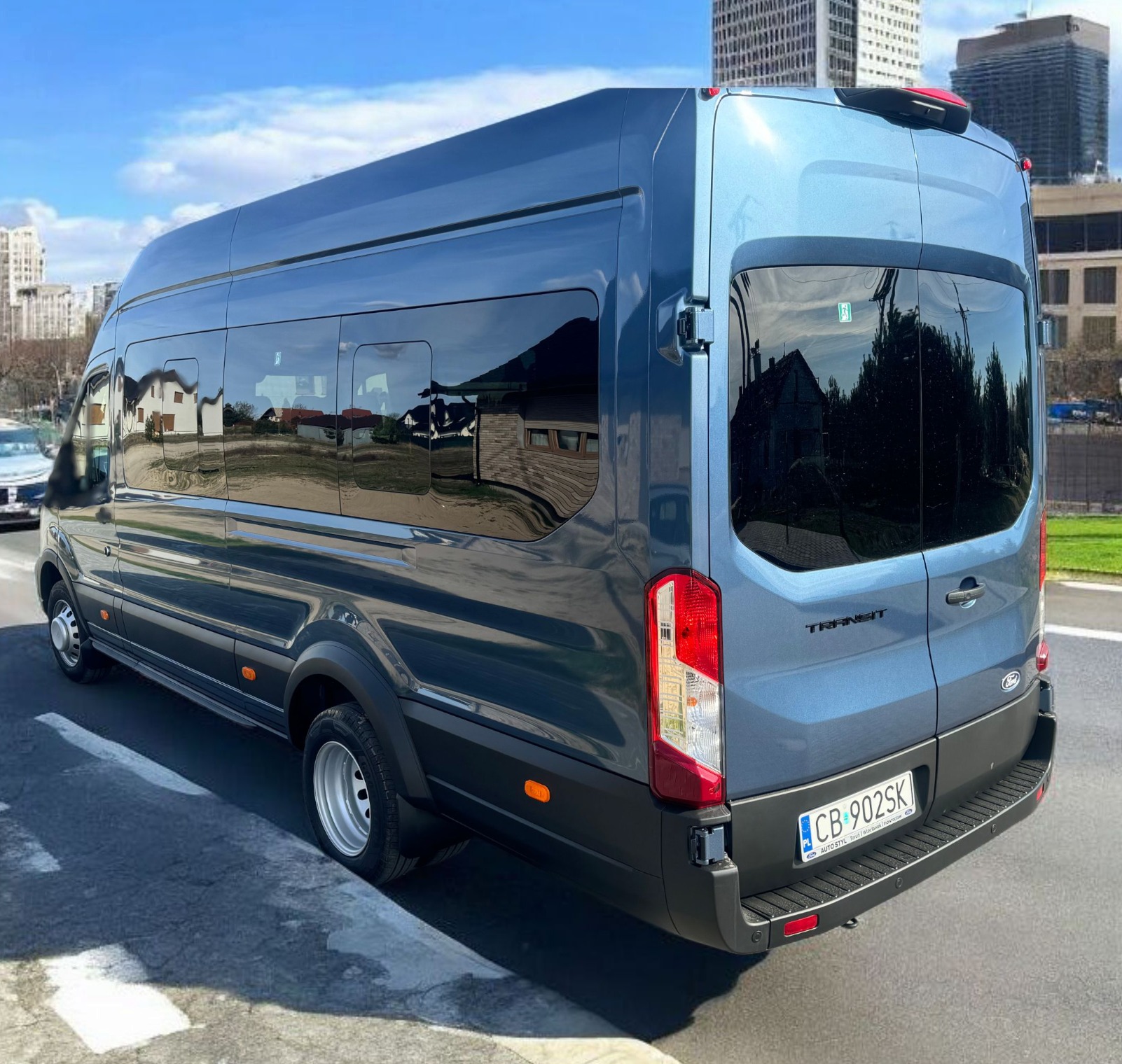 Niebieski bus Ford Transit z ciemnymi szybami, zaparkowany na tle miejskiego krajobrazu z budynkami. Widoczny tył pojazdu i tablica rejestracyjna.