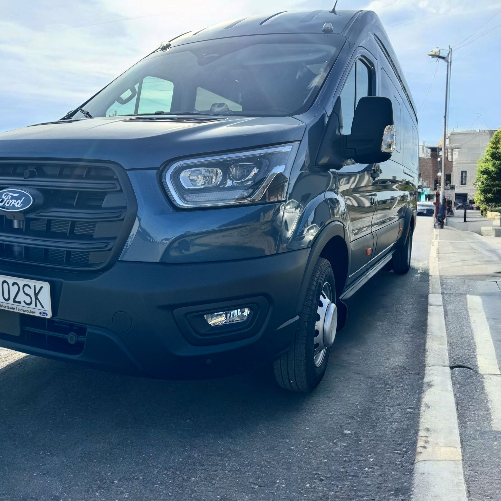Niebieski Ford Transit z widoczną tablicą rejestracyjną zaparkowany na ulicy w słoneczny dzień. Widok z przodu, lekko z dołu.