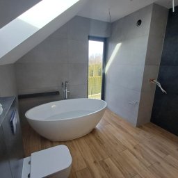 Damian Niedziałek - Nowoczesna łazienka na poddaszu z wolnostojącą wanną, drewnopodobną podłogą i oknem dachowym. Szare płytki na ścianach i czarna ściana z instalacją elektryczną.