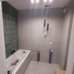 Damian Niedziałek - Nowoczesna łazienka w trakcie wykańczania: szare płytki, zielona mozaika nad wanną, minimalistyczna lampa wisząca i widoczne rury instalacyjne.