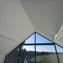 Damian Niedziałek - Wnętrze z oknem o trójkątnym szczycie i czarnych ramach, widok na zielony krajobraz. Białe ściany i sufit, nowoczesny design.