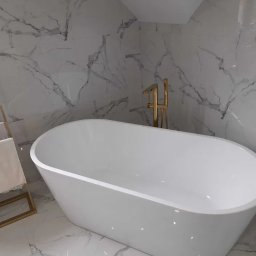 Damian Niedziałek - Nowoczesna, wolnostojąca wanna owalna z złotą armaturą na tle marmurowej ściany. Obok złoty wieszak na ręczniki. Minimalistyczny design.