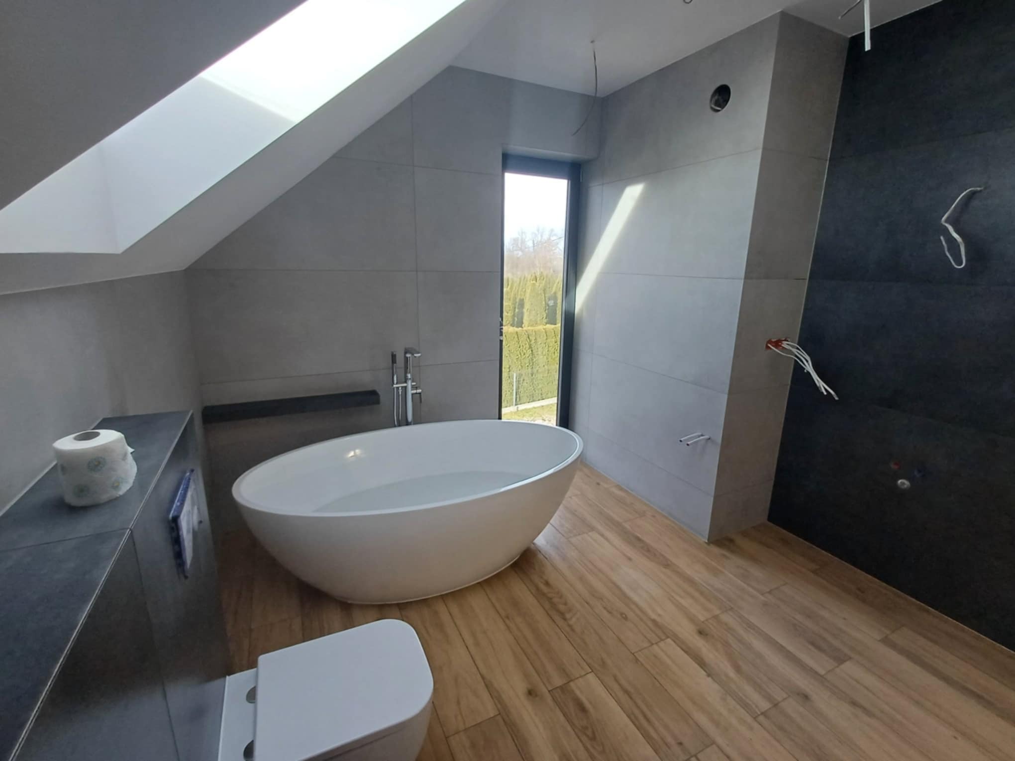 Nowoczesna łazienka na poddaszu z wolnostojącą wanną, drewnopodobną podłogą i oknem dachowym. Szare płytki na ścianach i czarna ściana z instalacją elektryczną.