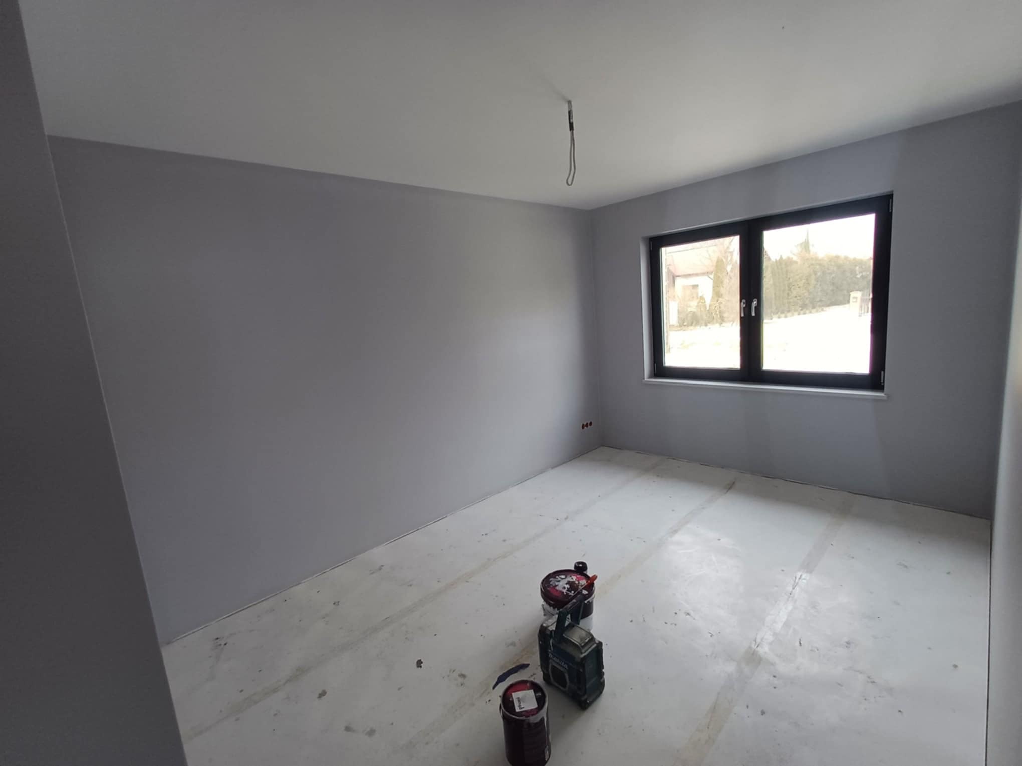 Pomieszczenie po malowaniu ścian na szaro, widoczne narzędzia malarskie i okno z czarną ramą. Podłoga przygotowana do dalszych prac wykończeniowych.