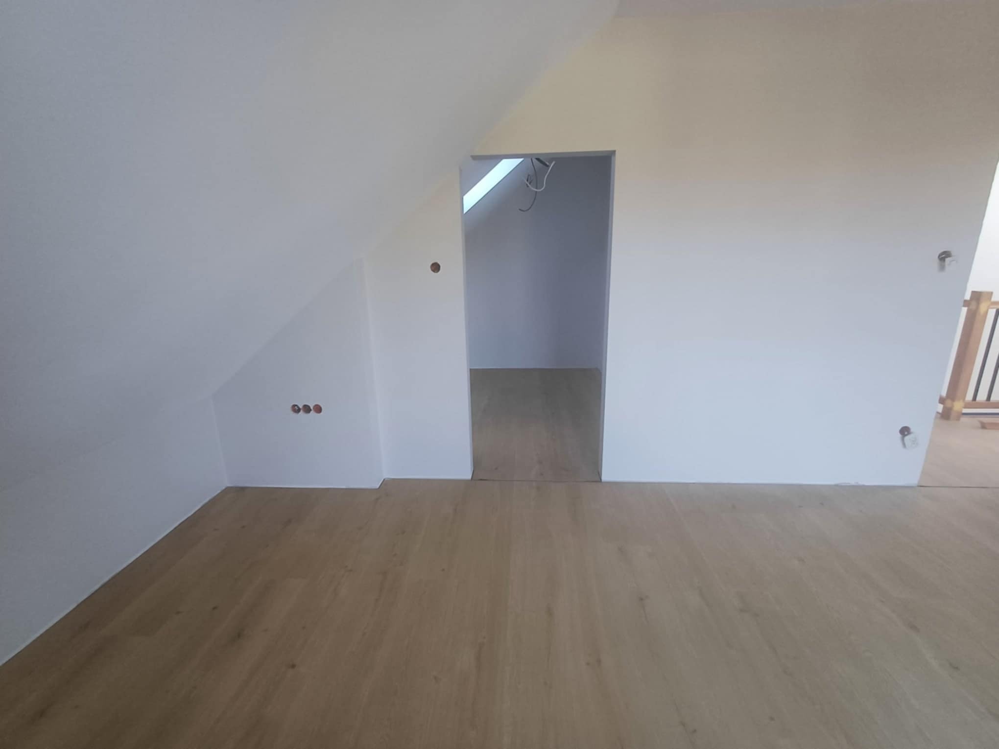 Jasne panele podłogowe w pokoju na poddaszu z białymi ścianami. Widoczne gniazdka i wejście do innego pomieszczenia. Ukończone wykończenie poddasza.