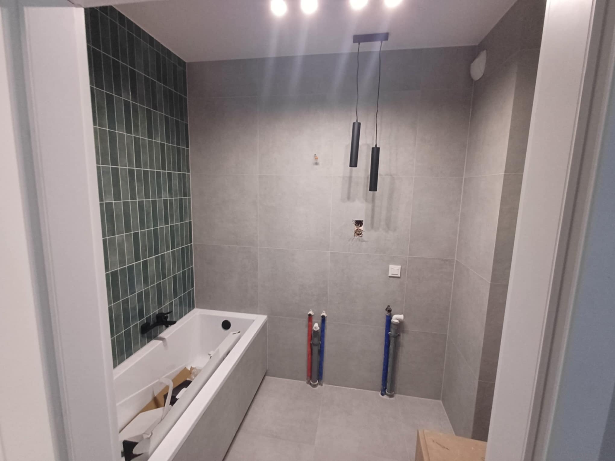 Nowoczesna łazienka w trakcie wykańczania: szare płytki, zielona mozaika nad wanną, minimalistyczna lampa wisząca i widoczne rury instalacyjne.