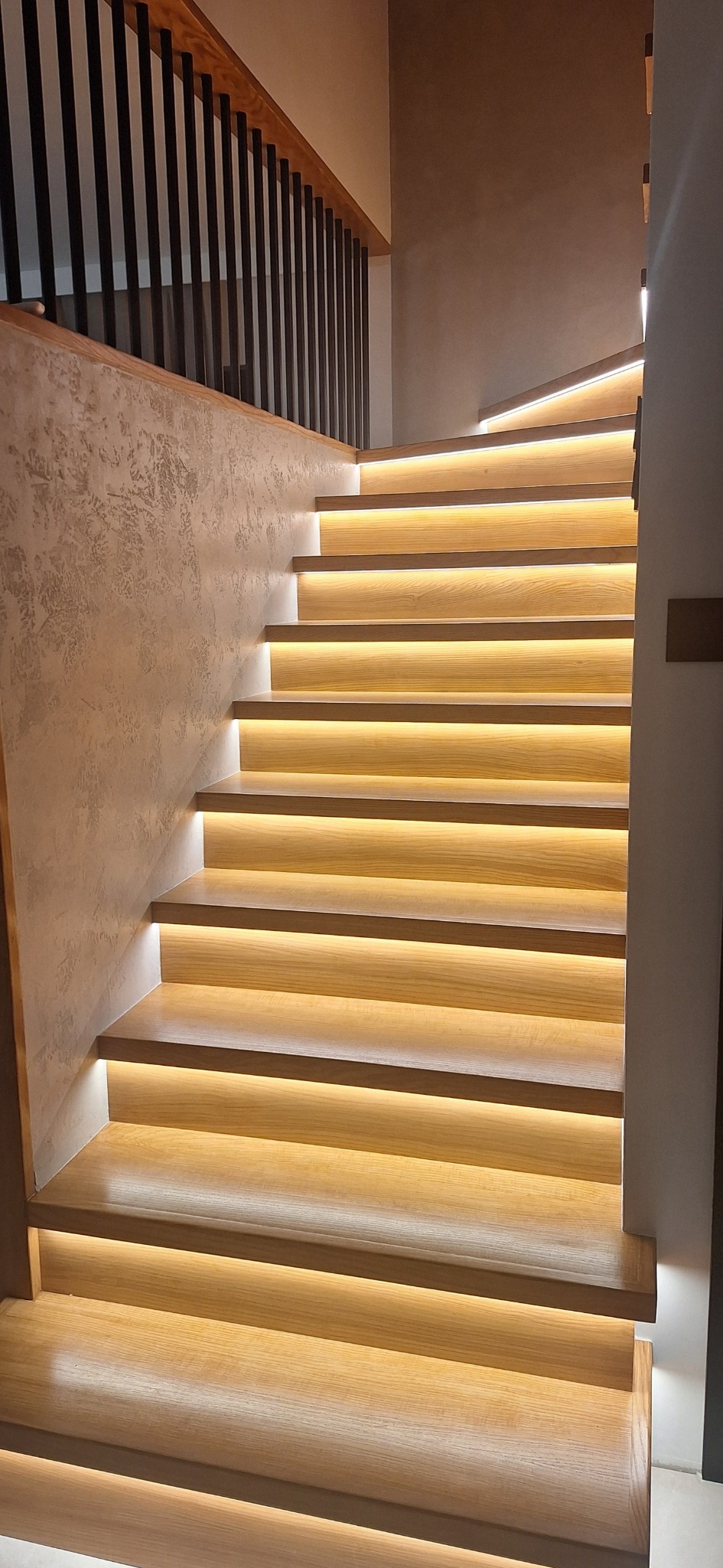 Nowoczesne schody drewniane z podświetleniem LED, balustrada z czarnych prętów. Ściana wykończona dekoracyjnym tynkiem. Oświetlenie tworzy ciepły, nastrojowy klimat.