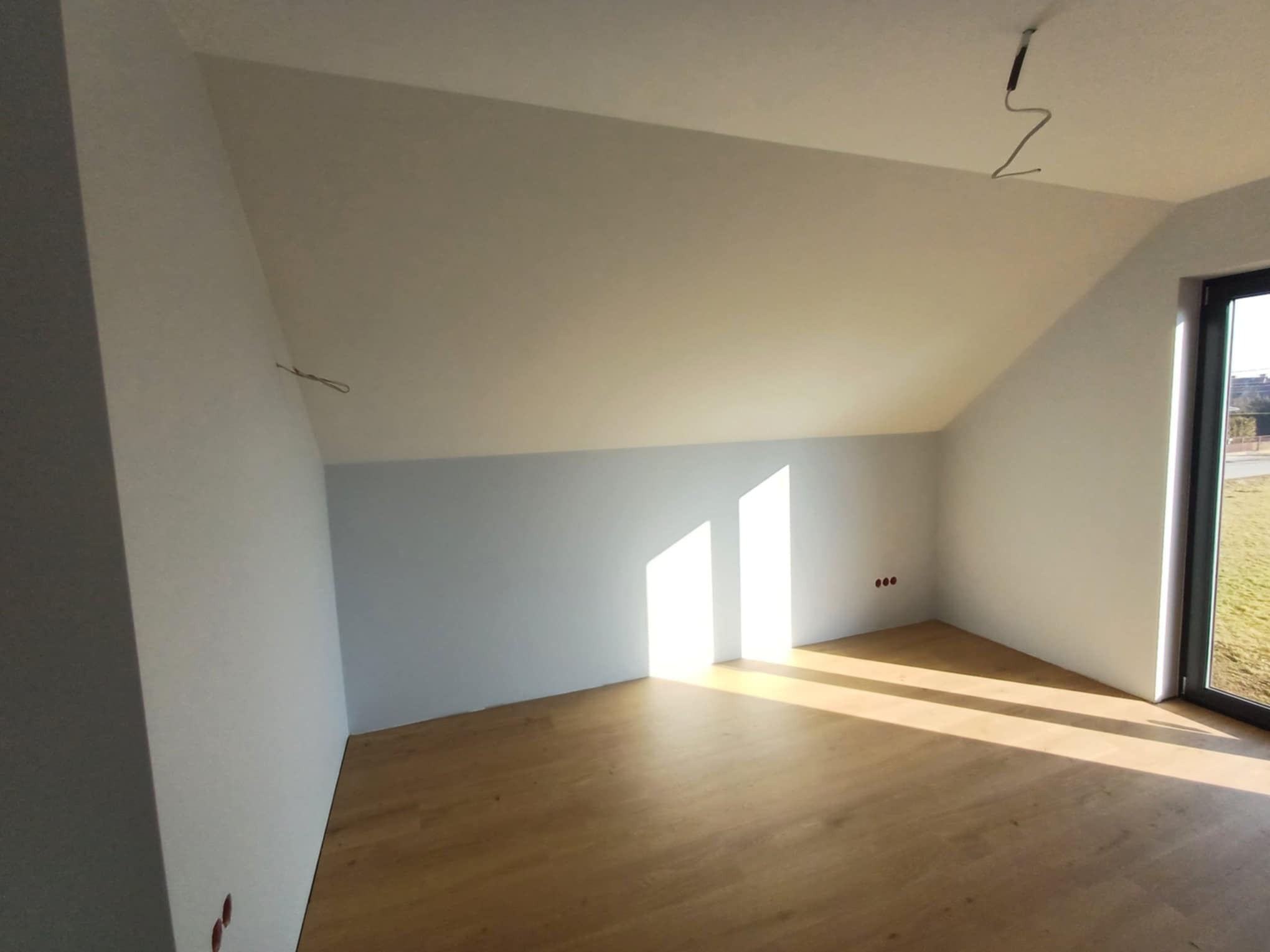 Jasne poddasze po remoncie: świeżo pomalowane ściany w bieli i błękicie, drewniana podłoga, okno z widokiem na zieleń, kabel zwisający z sufitu. Promienie słoneczne oświetlają wnętrze.