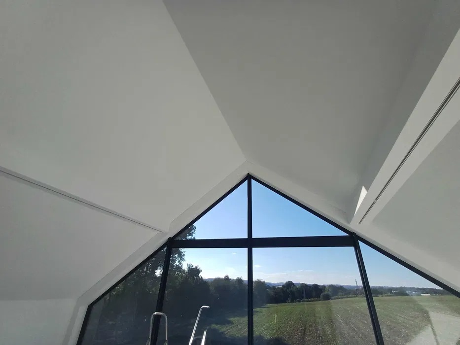 Wnętrze z oknem o trójkątnym szczycie i czarnych ramach, widok na zielony krajobraz. Białe ściany i sufit, nowoczesny design.