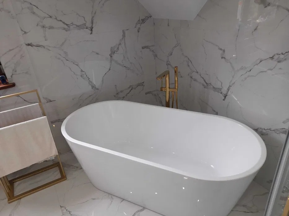 Nowoczesna, wolnostojąca wanna owalna z złotą armaturą na tle marmurowej ściany. Obok złoty wieszak na ręczniki. Minimalistyczny design.