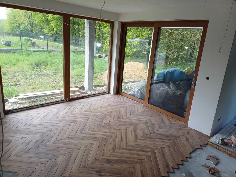 Układanie parkietu w jodełkę w jasnym pokoju z dużymi oknami i przesuwnymi drzwiami balkonowymi. Widoczne niedokończone prace przy ścianie.