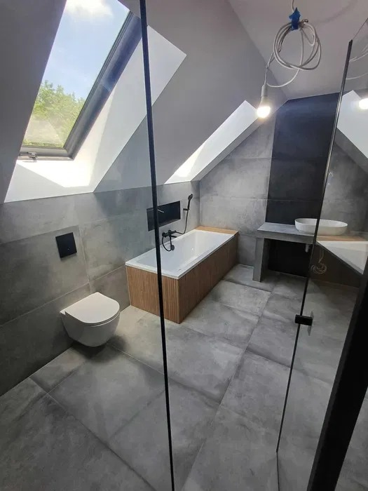 Nowoczesna łazienka na poddaszu z oknem dachowym. Szare płytki, biała wisząca toaleta i wanna z drewnianą obudową. Kabina prysznicowa ze szklanymi drzwiami.