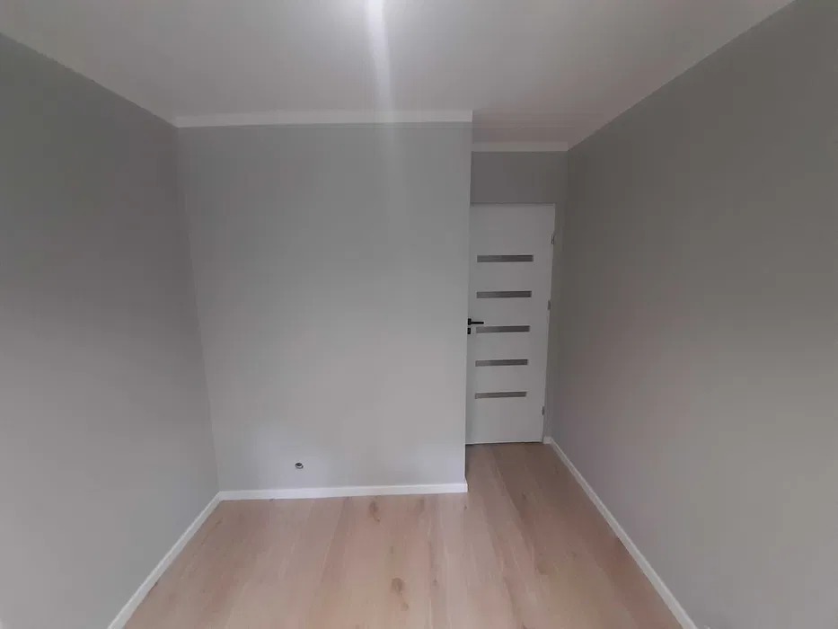 Wnętrze pokoju po remoncie: świeżo pomalowane szare ściany z białą listwą przypodłogową, jasna drewniana podłoga i białe drzwi z poziomymi przeszkleniami.