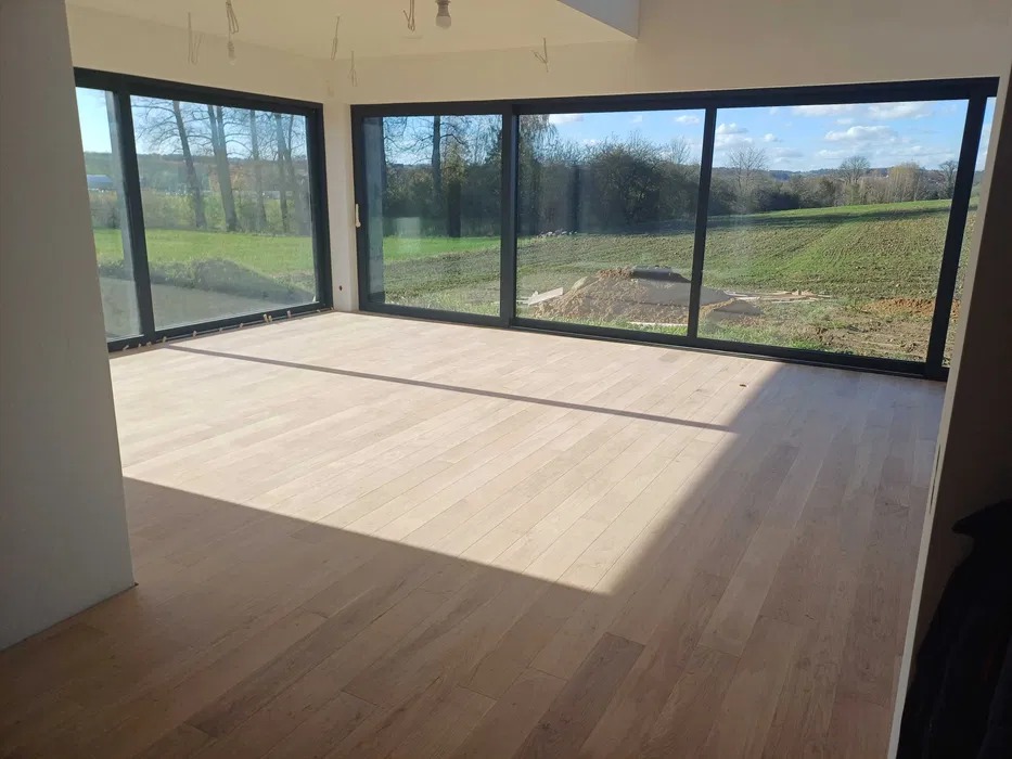 Jasna drewniana podłoga w przestronnym pokoju z dużymi oknami. Widok na zielony krajobraz za oknem. Świeże wykończenie, naturalne światło.